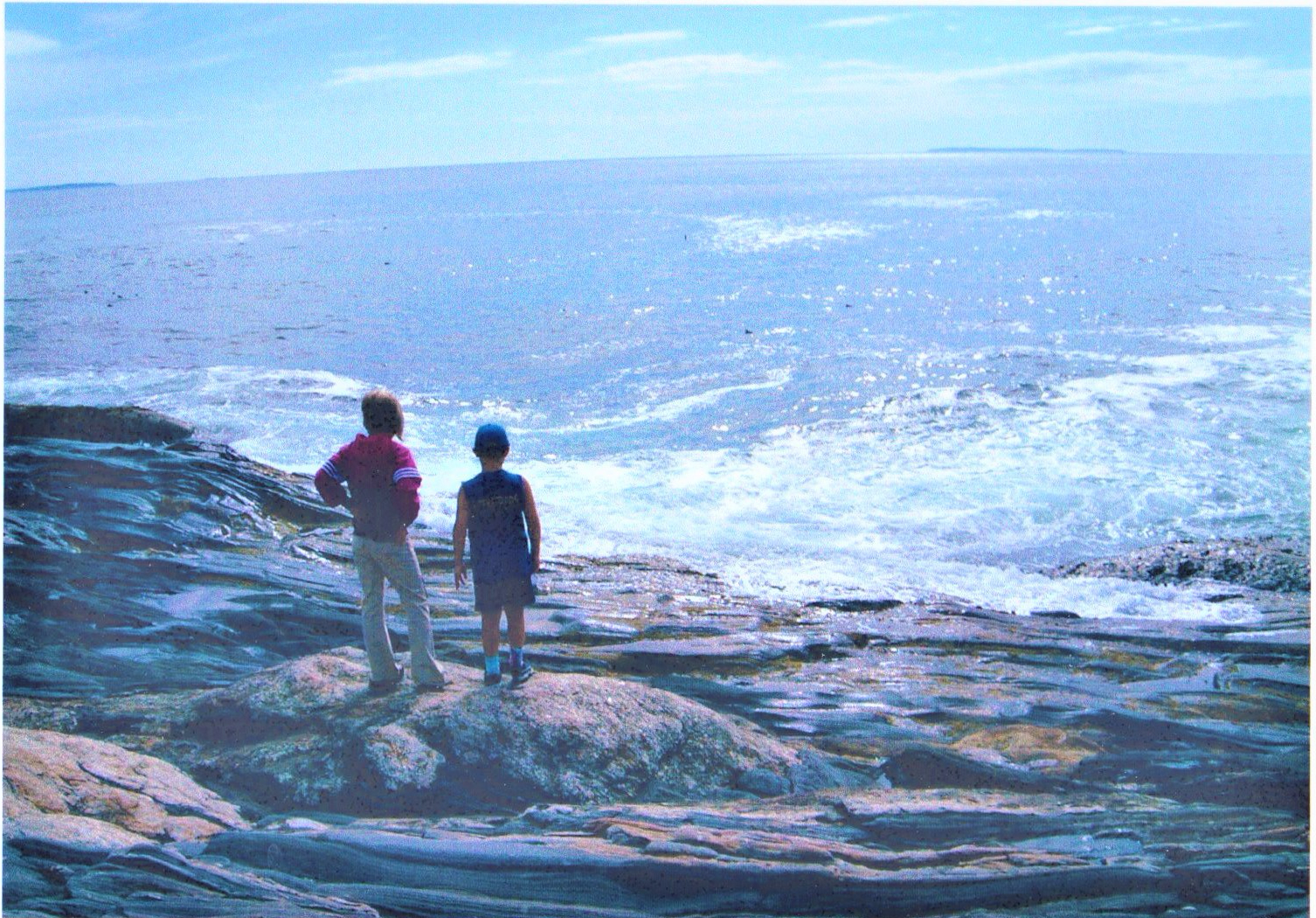 Sister and brother look out at the sparking blue ocean from atop a weathered rock.