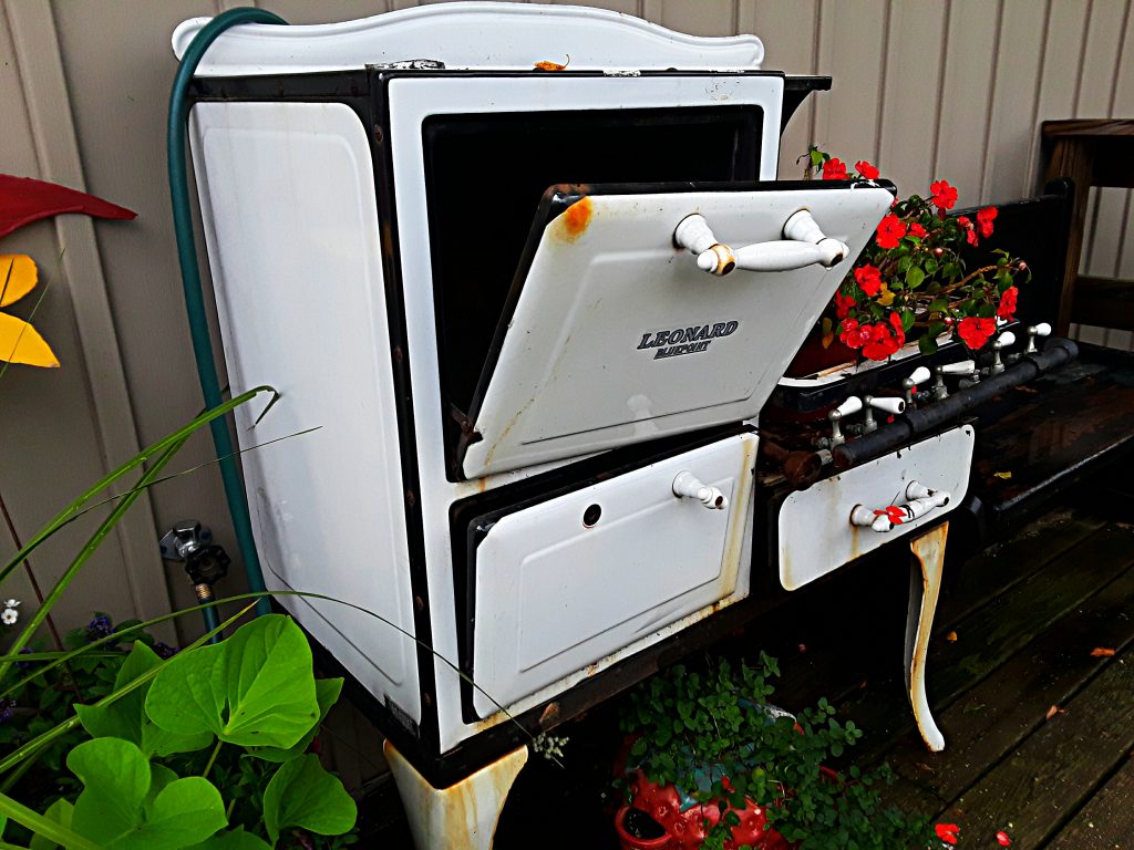An old white enamel oven is planted with red impatiens outside the market door.